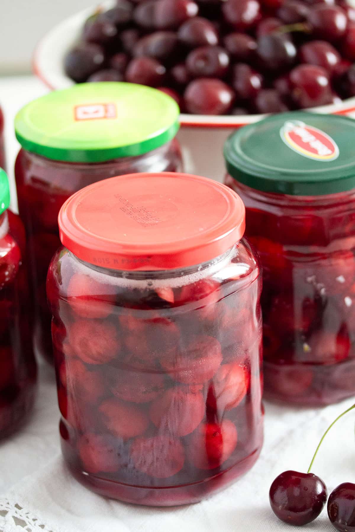 three jars with preserved cherries and a bowl of fresh cherries behind them.