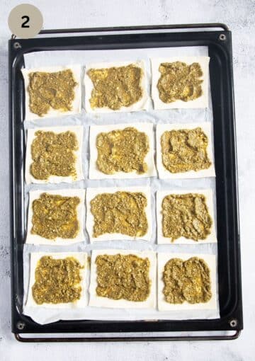 twelve puff pastry squares smeared with pesto on a baking sheet.