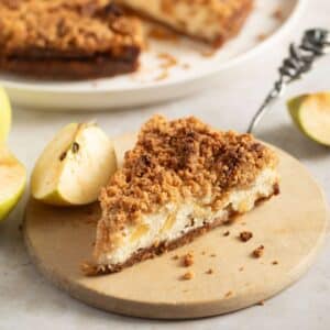 apple streusel cheesecake slice on a small round wooden board and apple pieces around it.