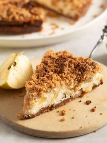 apple streusel cheesecake slice on a small round wooden board and apple pieces around it.