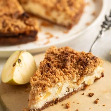 slice of apple cheesecake with streusel, the whole cake behind it on a platter.