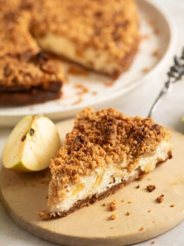 slice of apple cheesecake with streusel, the whole cake behind it on a platter.