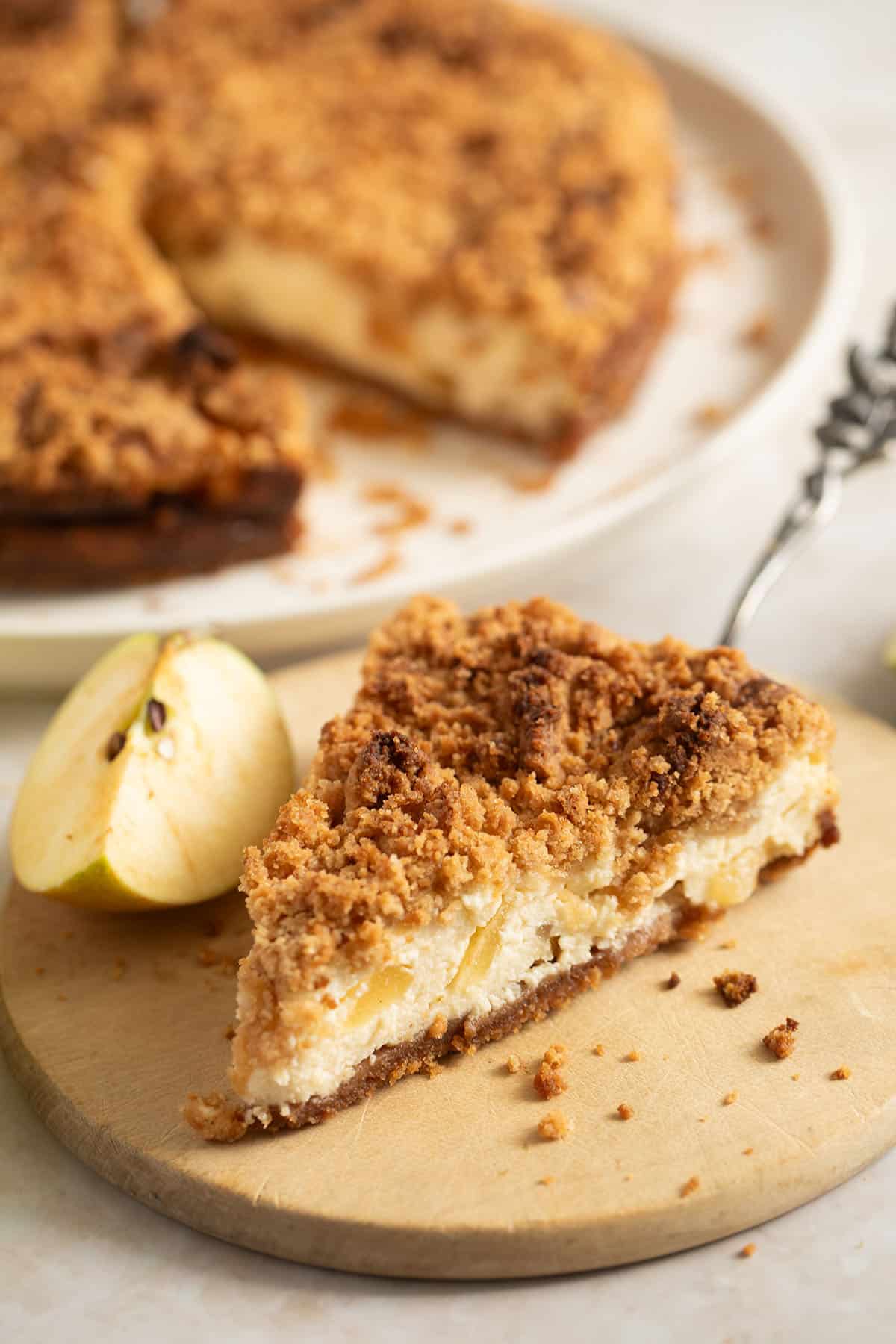 slice of apple cheesecake with streusel, the whole cake behind it on a platter.