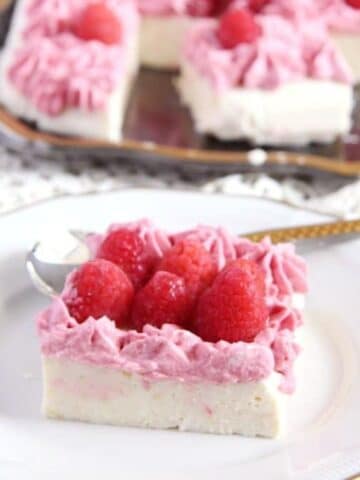 a square piece of ricotta cheesecake with fresh raspberries on top, the rest of the cake behind it.