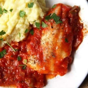 close up of a piece of fish in tomato sauce served with mashed potatoes.