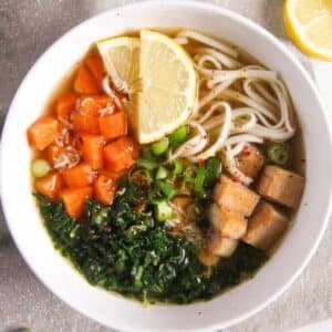 vegan tofu soba noodle soup with kale and lemon wedges in a bowl.