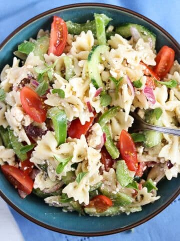 greek-style potluck pasta salad with tomatoes, cucumbers, and feta in a bowl on a blue kitchen cloth.
