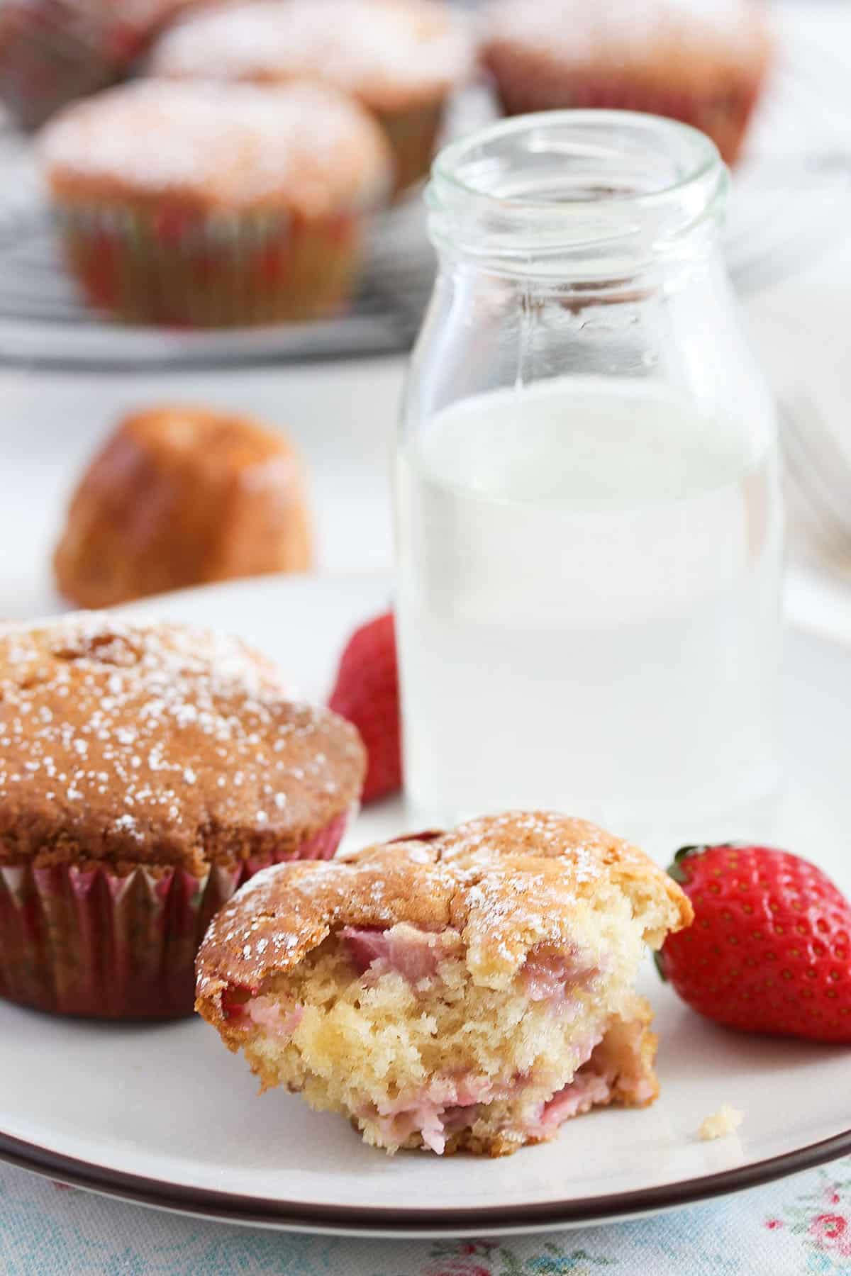 one split muffin showing the strawberry pieces inside, a milk bottle and more muffins behind it.