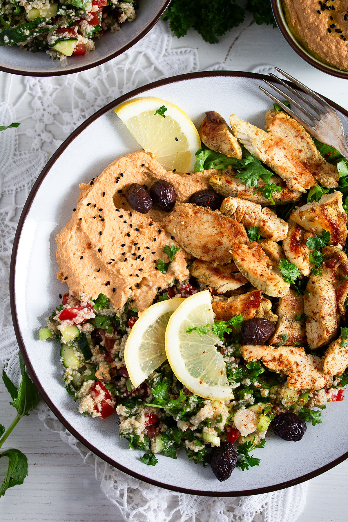 overhead view of a platter with chicken strips, hummus, olives, tabbouleh, and lemon wedges.