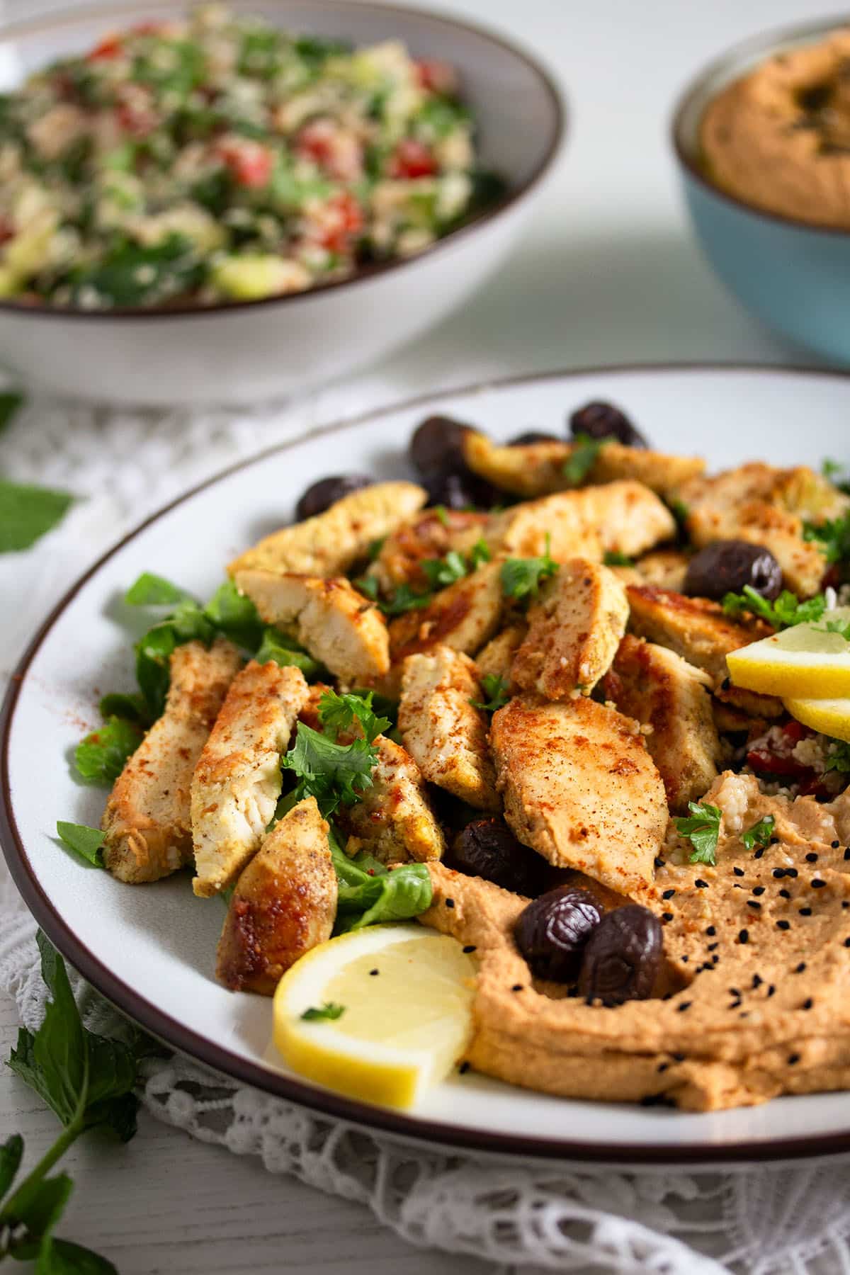 golden chicken breast pieces served with hummus, olives and lemon with a bowl of tabbouleh behind it.