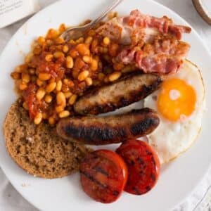 full english breakfast with beans, sausages, egg, bread, bacon, and grilled tomatoes on a plate.