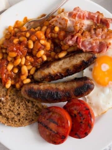 full english breakfast with beans, sausages, egg, bread, bacon, and grilled tomatoes on a plate.