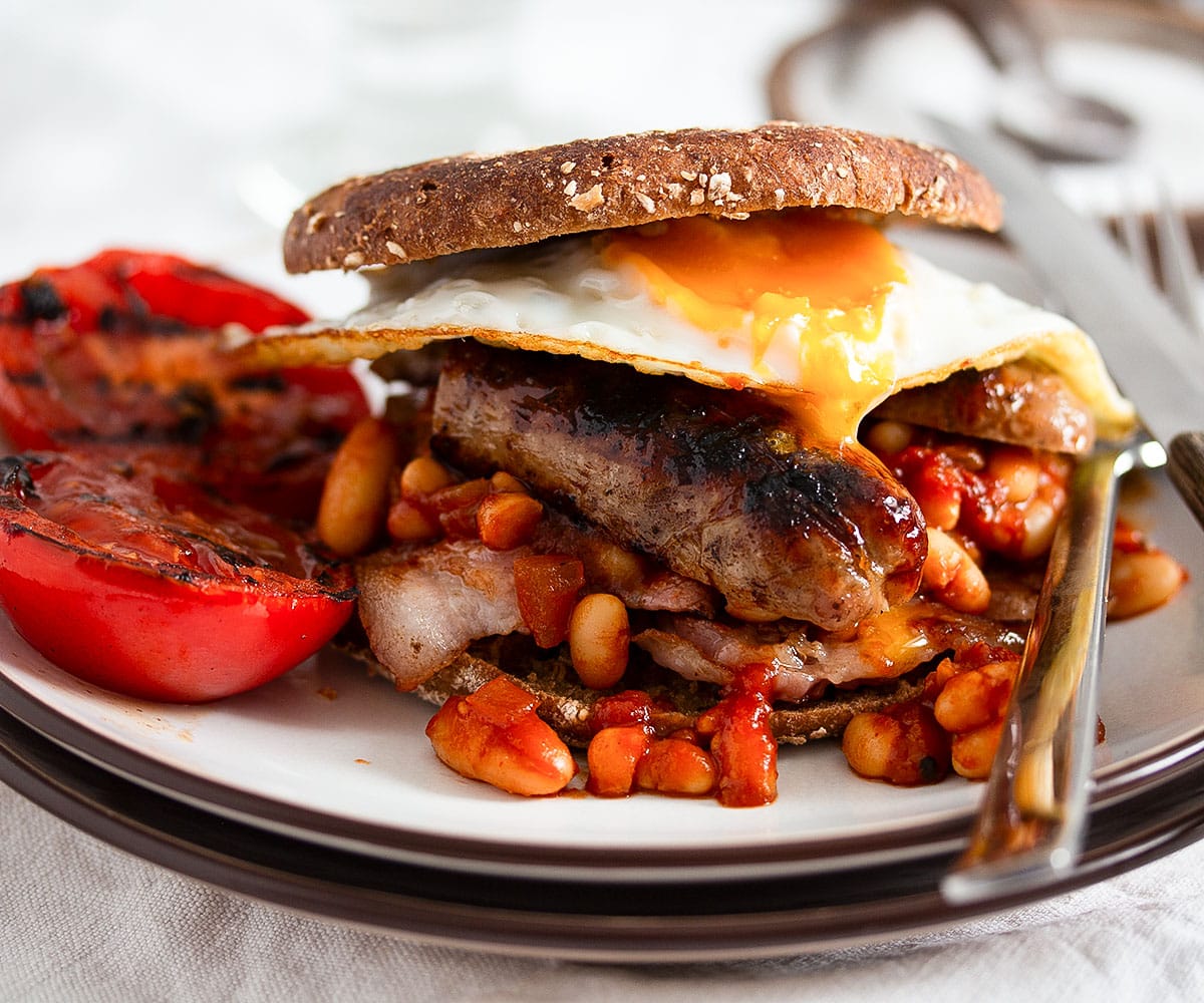 english breakfast with beans served in a toasted muffin with fried tomatoes on the side.