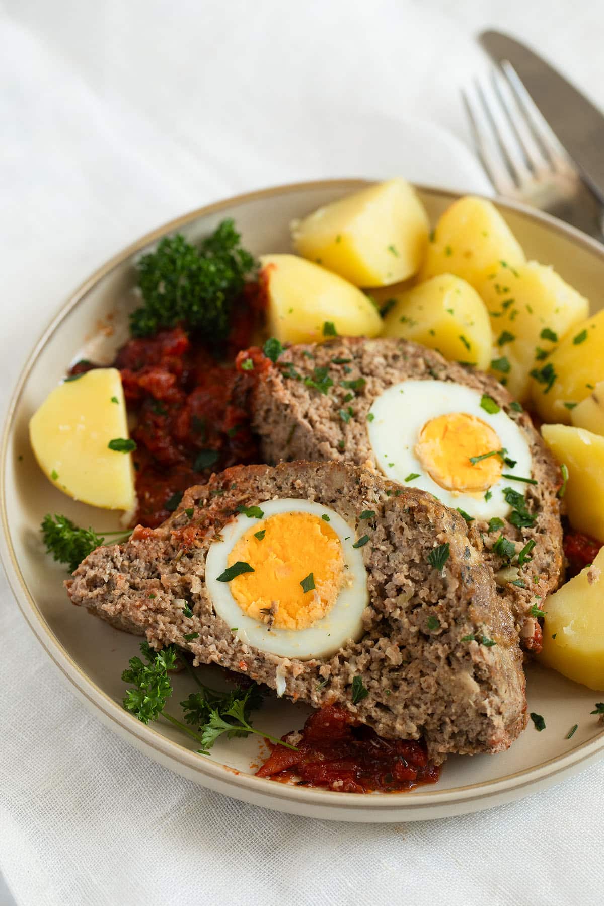 two thick slices of meatloaf with egg inside served with boiled potatoes and tomato sauce on a plate.