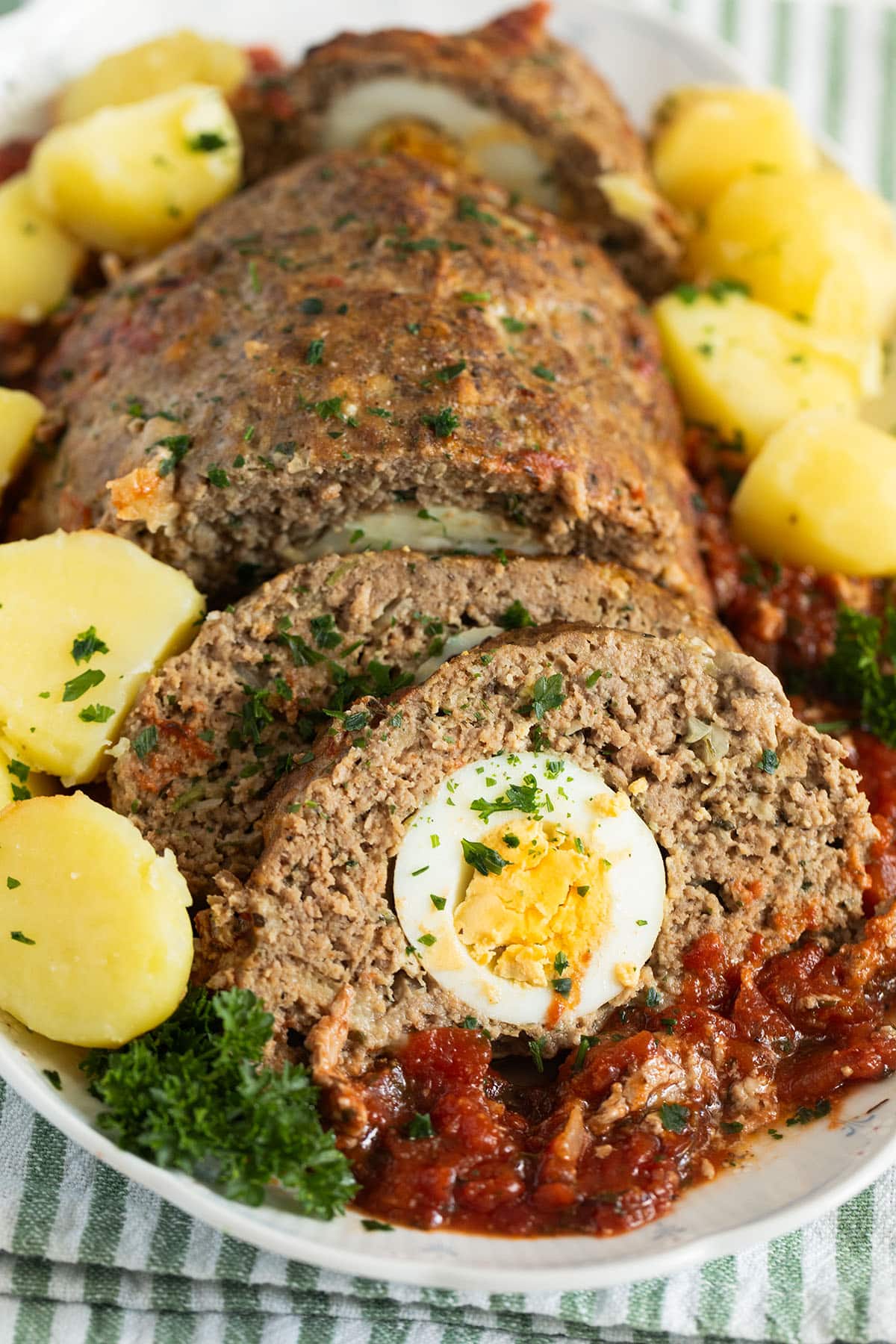 sliced meatloaf with eggs inside ona platter with boiled potatoes and tomato sauce around it.
