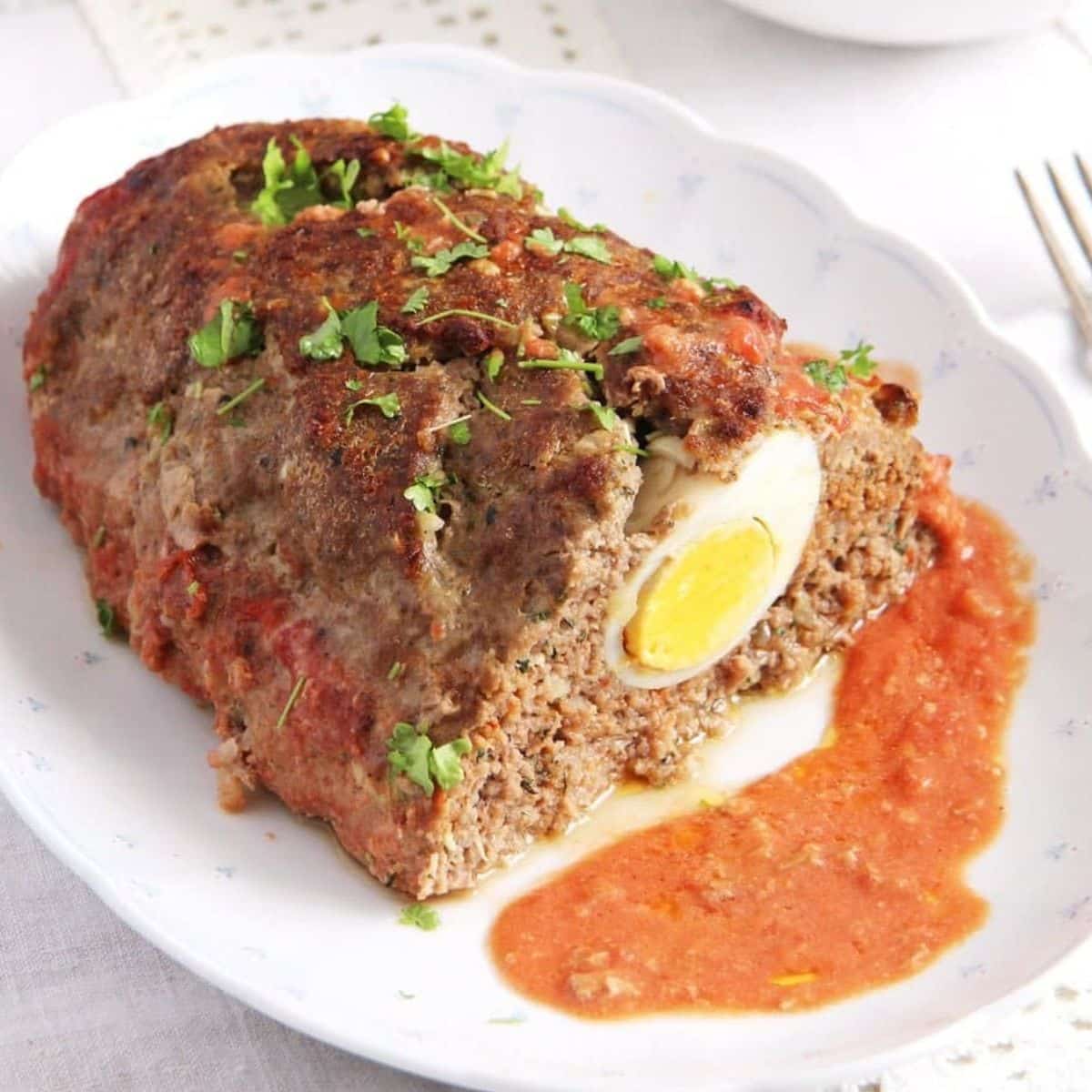 meatloaf with eggs cut to show the egg inside with sauce on a platter.