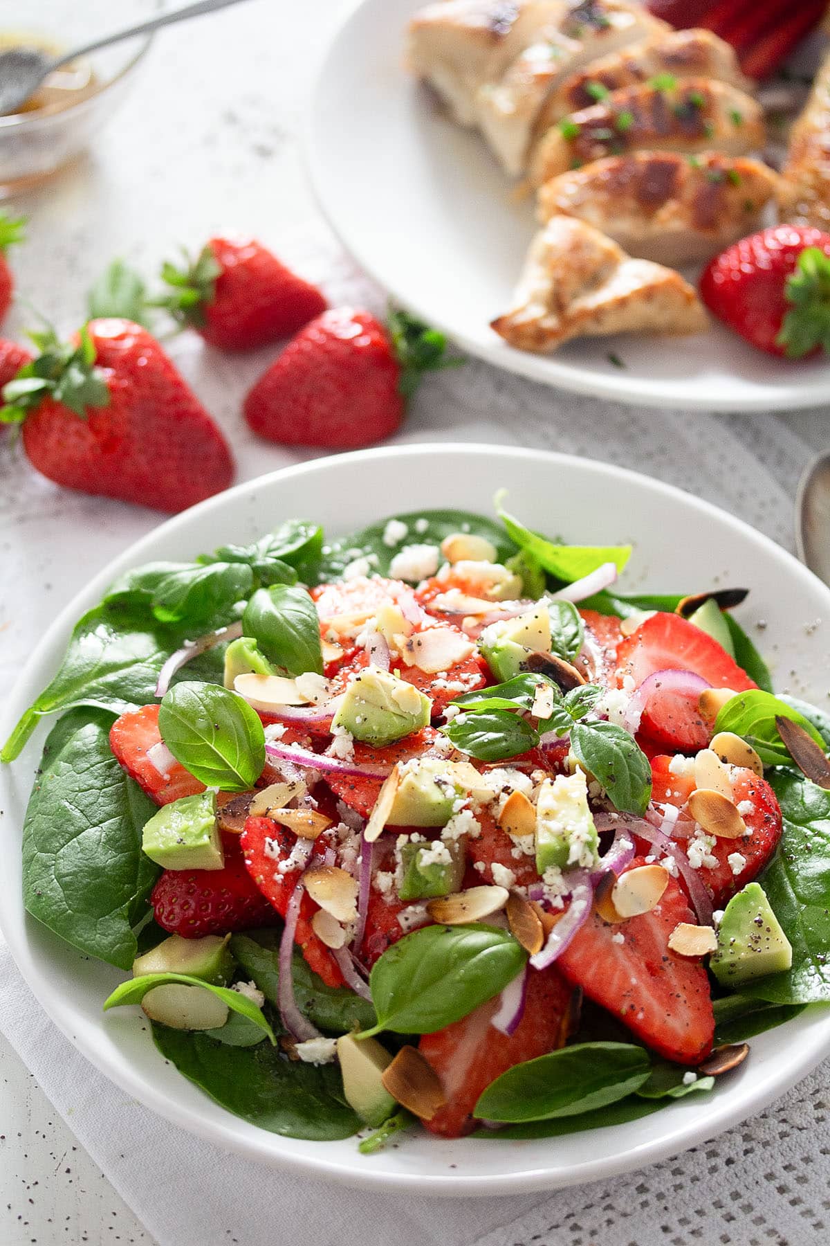 strawberry salad with goat cheese and almonds on a plate, fresh strawberries and grilled chicken breast behind it.
