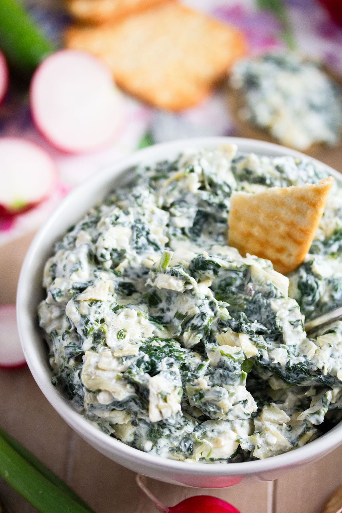 cold spinach dip with artichokes in a bowl with a cracker in it. 