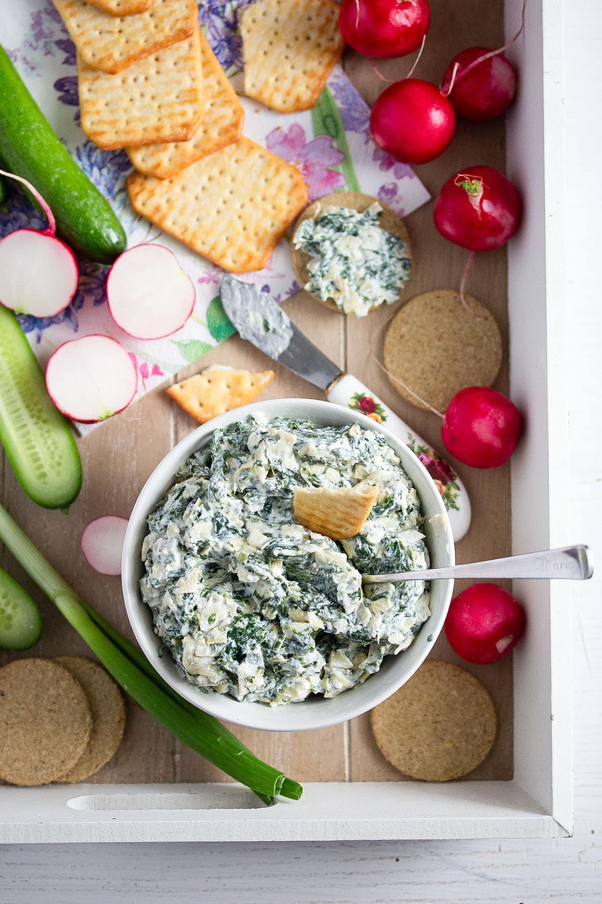 platter with spinach artichoke dip in a bowl served with crackers, mini cucumbers and radishes.