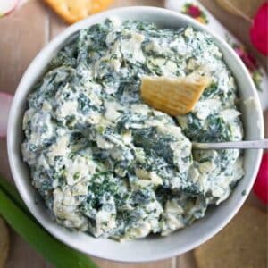 no-bake spinach artichoke dip in a bowl with a cracker and a spoon in it.