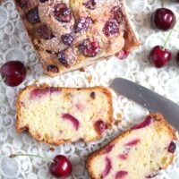 cherry loaf cake sliced on a vintage cloth with a knife and fresh fruit around it.