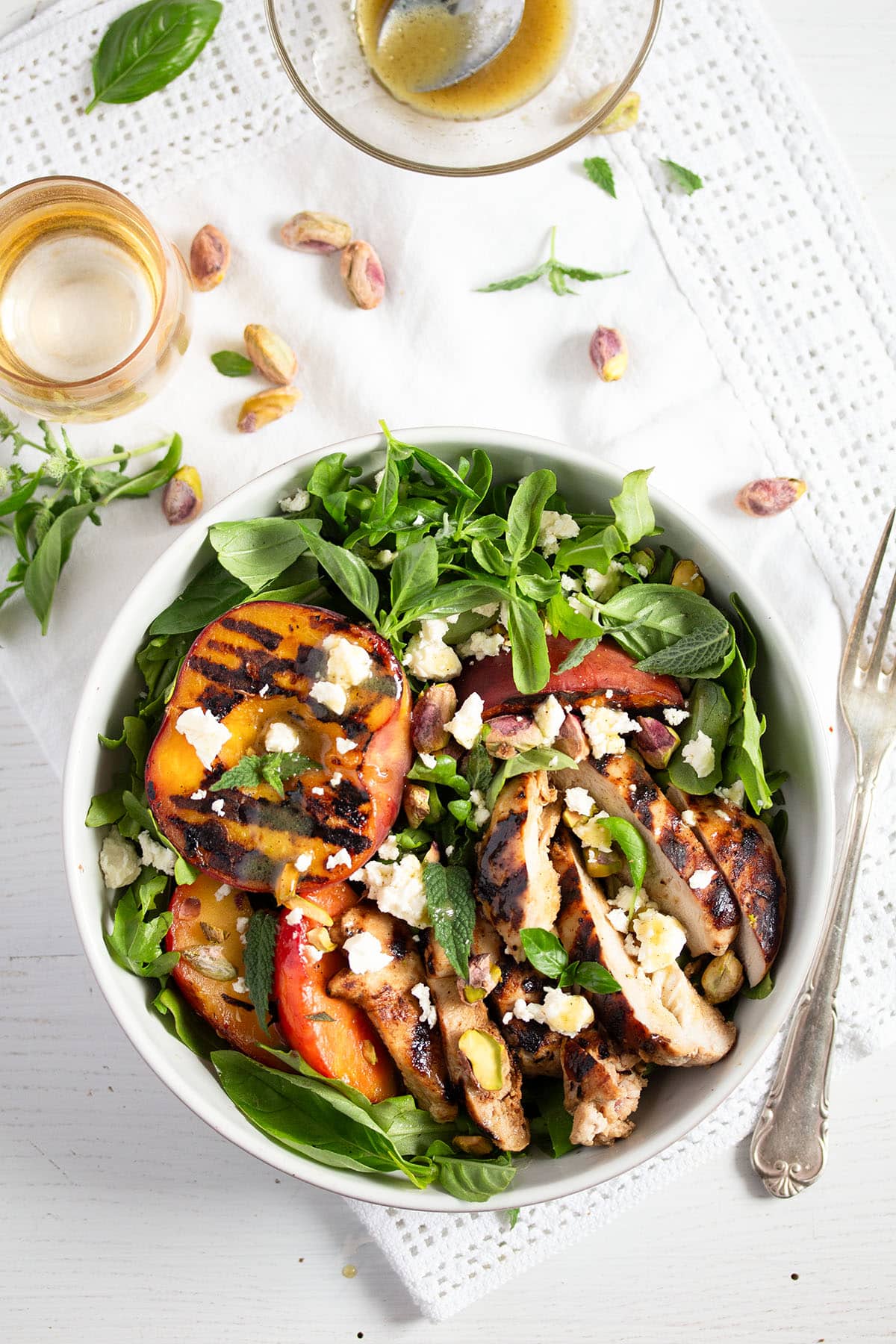 a bowl of peach and chicken breast salad with a glass, herbs and pistachios around it.