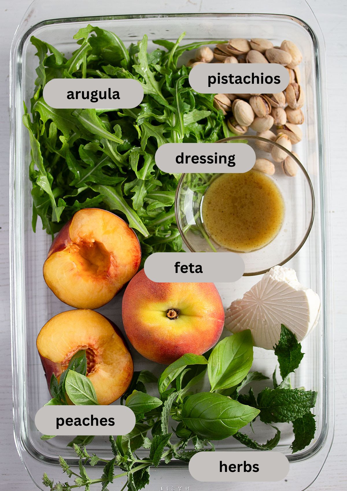 labeled ingredients for making peach salad with arugula and herbs in a glass baking dish.