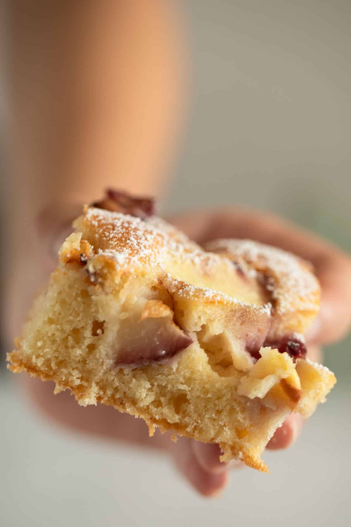 a hand holding a square piece of cake with peach pieces inside it.