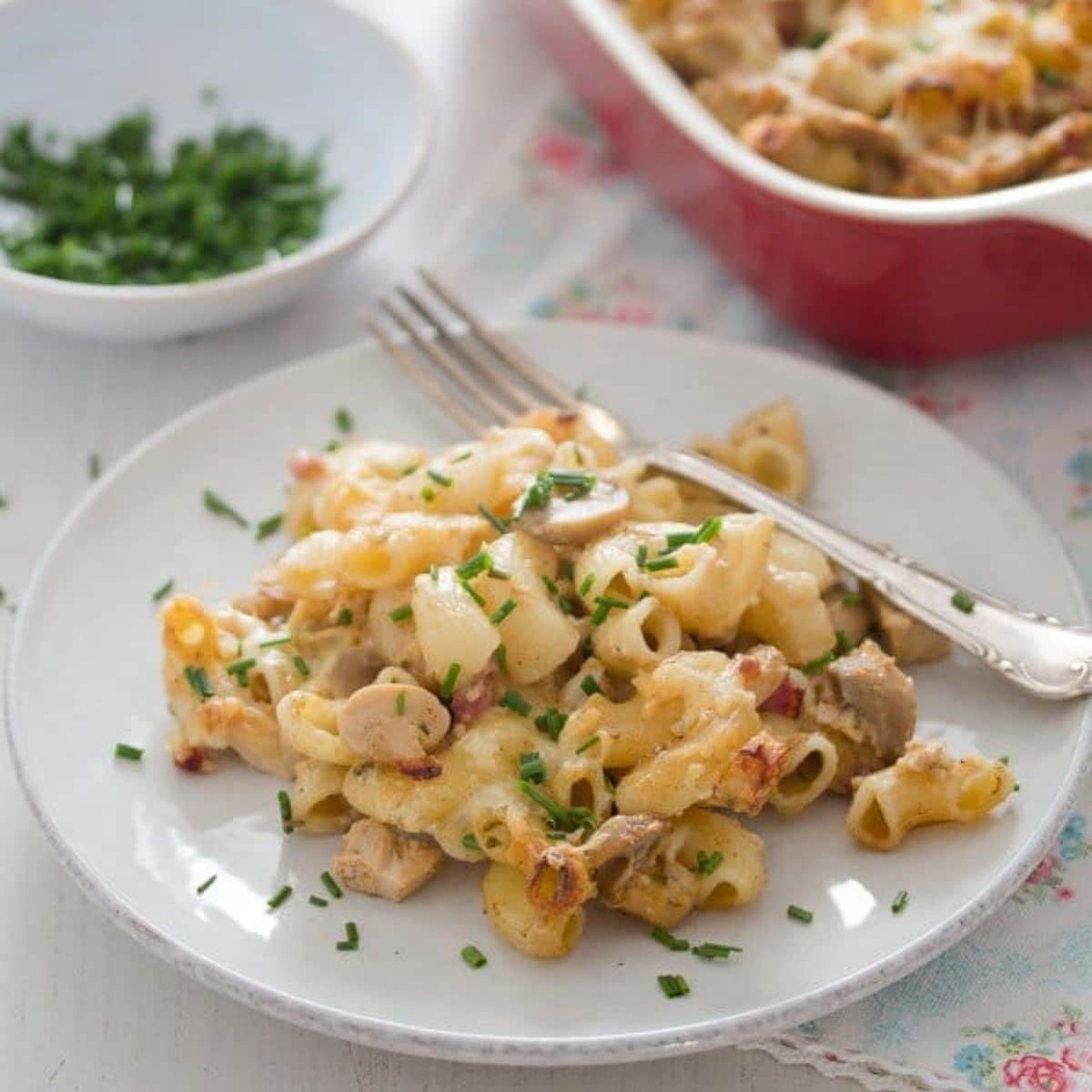 chicken, bacon and pasta bake on a small plate with a fork on it, a bowl of parsley behind it.
