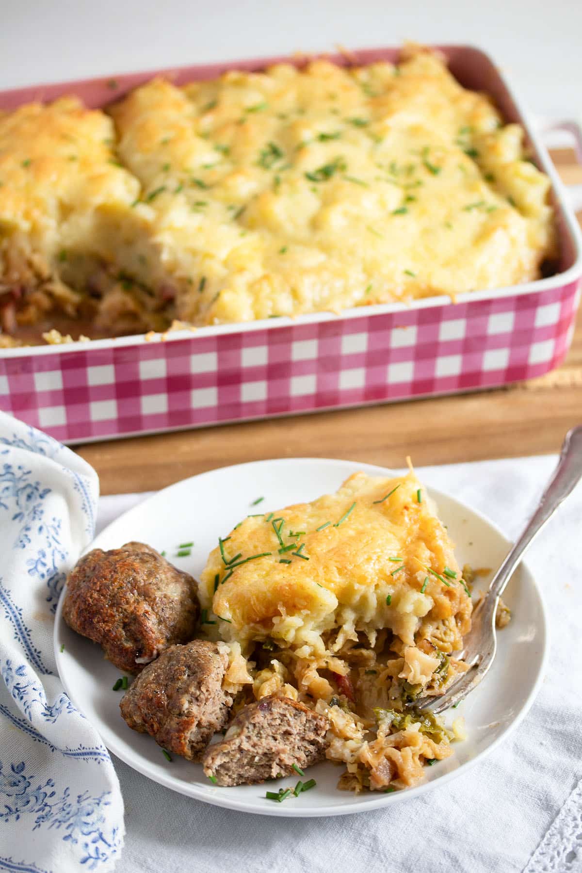 plate with cheesy baked cabbage and potatoes served with meatballs, the casserole dish behind it.