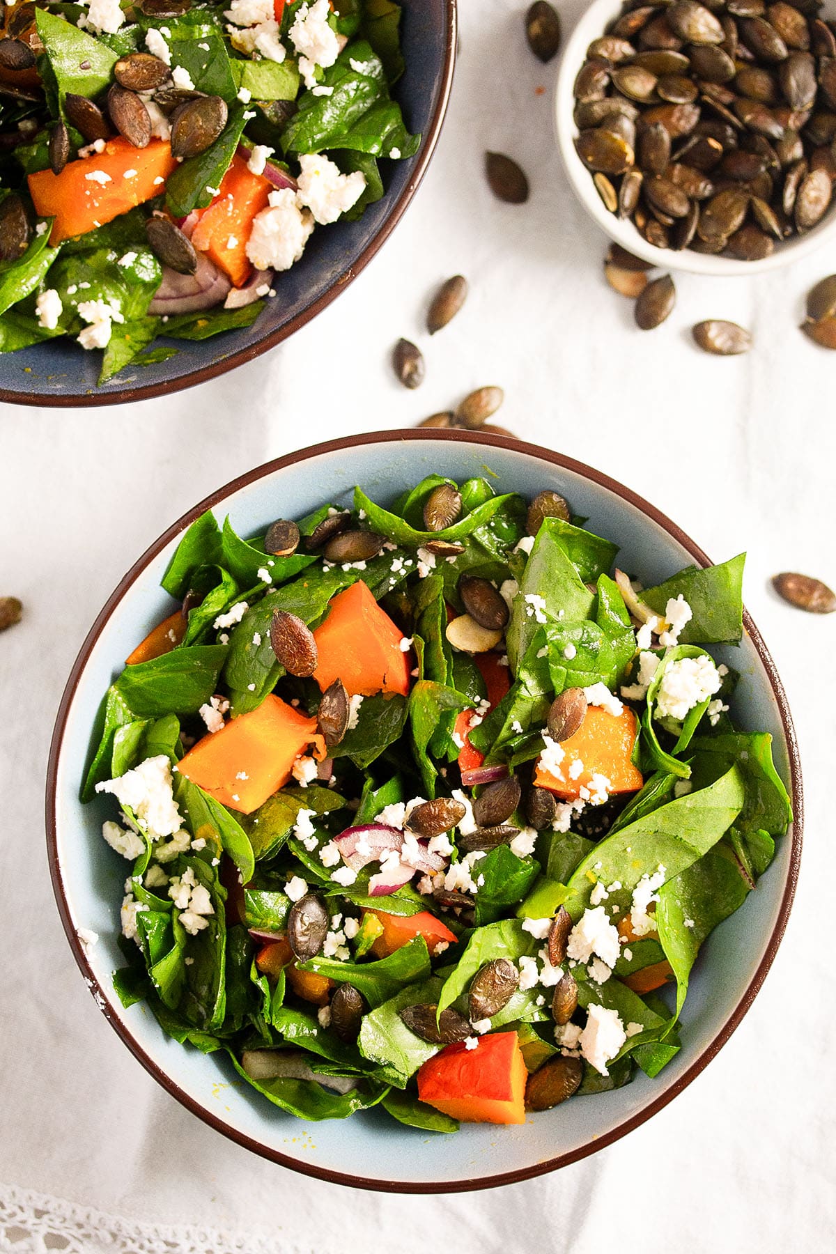 overhead view of two bowls of pumpkin salad with spinach and feta and a bowl of roasted pepitas.