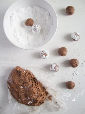 a piece of chocolate cookie dough, rolled cookie balls and some balls being rolled in a bowl of icing sugar.