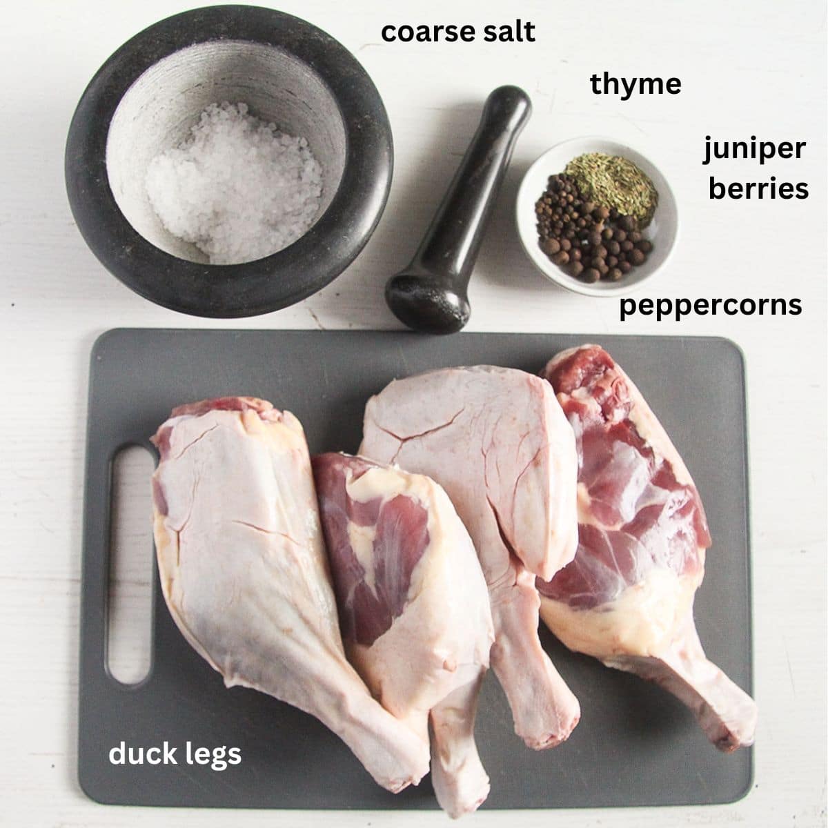 four raw duck legs, coarse salt in a mortar and spices in a bowl for making confit de canard recipe.