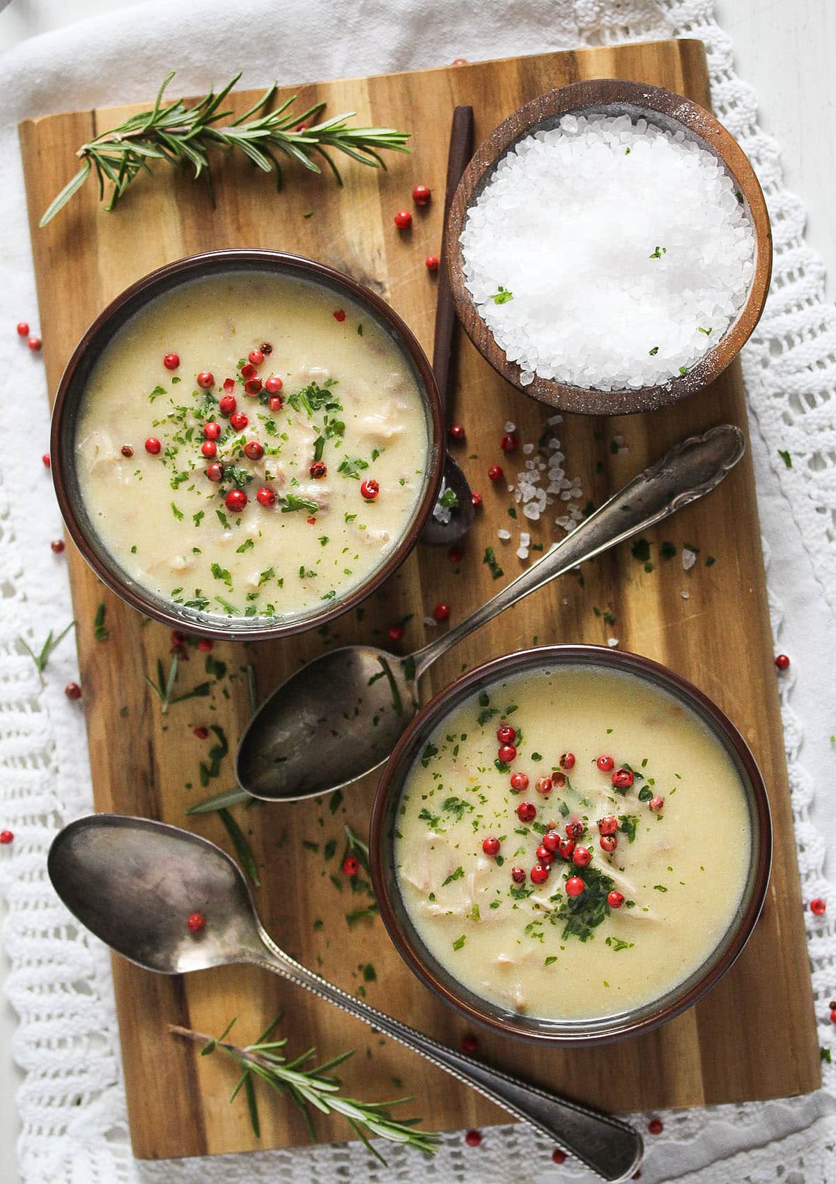 two bowls with turkey cream soup on a wooden board with two spoons, bowl of salt and rosemary sprigs.