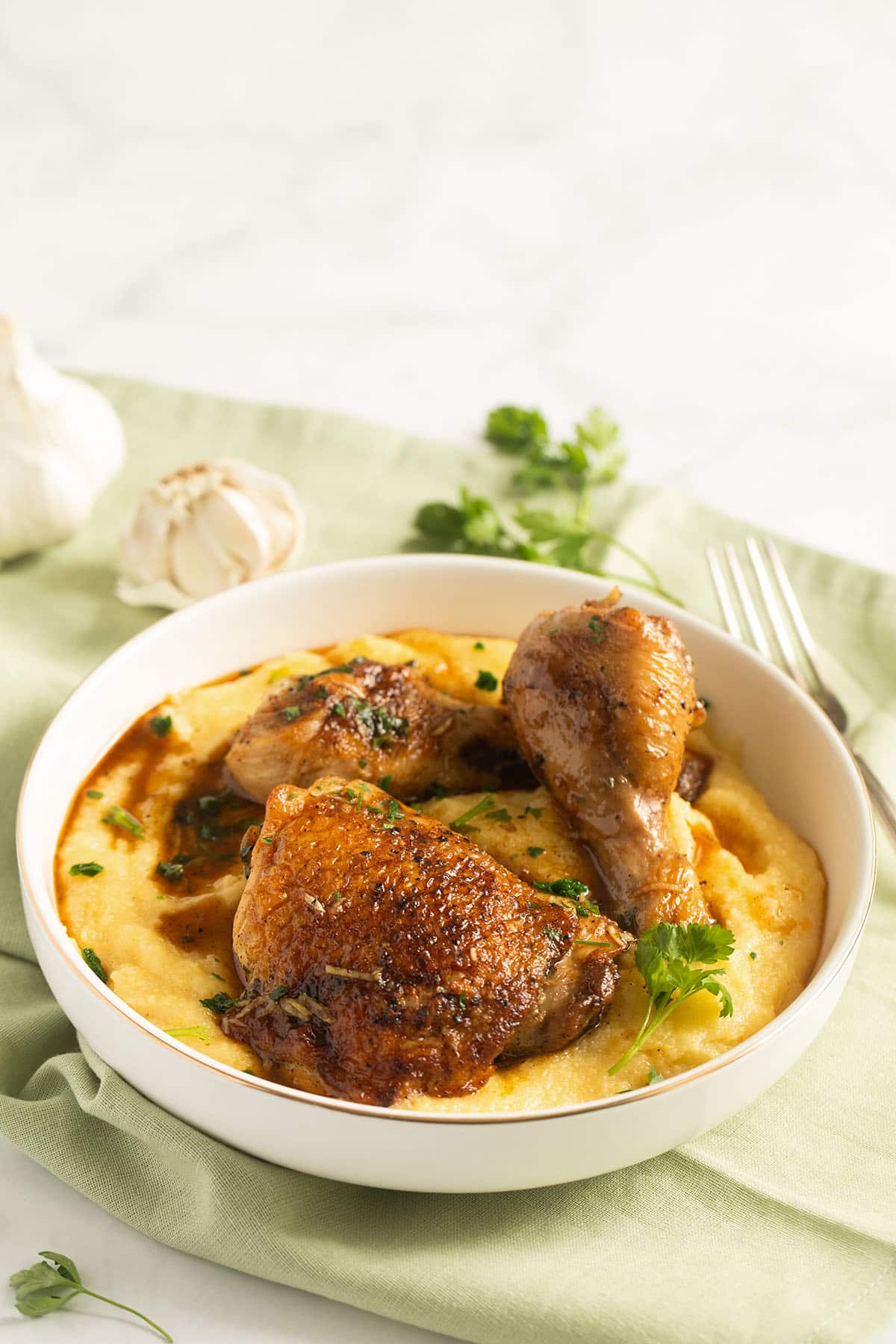a bowl with creamy polenta and chicken sprinkled with parsley, a garlic bulb and more parsley behind it.