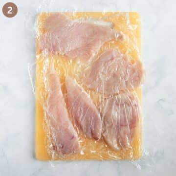 pounding chicken breast slices between layers of plastic wrap on a cutting board.