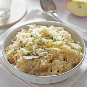 a small serving bowl full of cooked sauerkraut, half an apple, one spoon and a glass behind it.