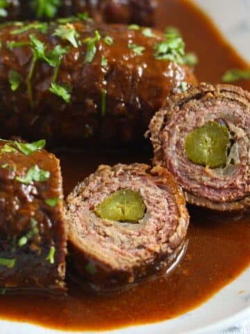 german beef roulades served in sauce and cut into several slices showing the gherkin inside.