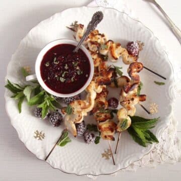 chicken skewers, a small cup full of blackberry sauce and mint leaves on a vintage plate.