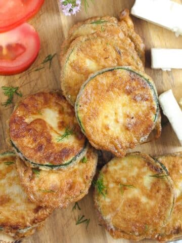 pan fried zucchini with flour on a wooden board with tomato and feta cheese slices.