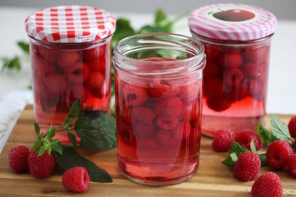 three jars of canned raspberries and fresh berries on a cutting board