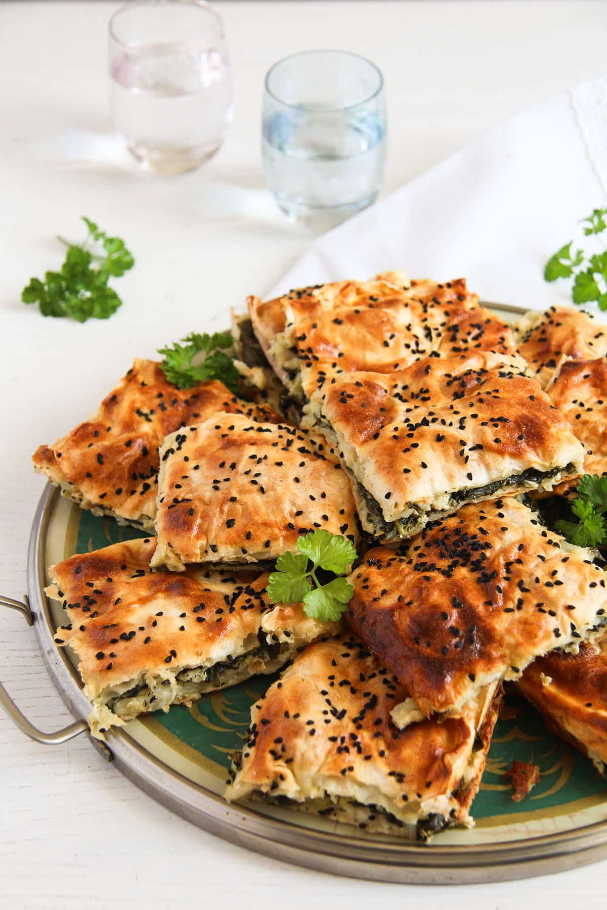 platter with stapled pieces of spinach borek sprinkled with nigella seeds and parsley, two small glasses behind.