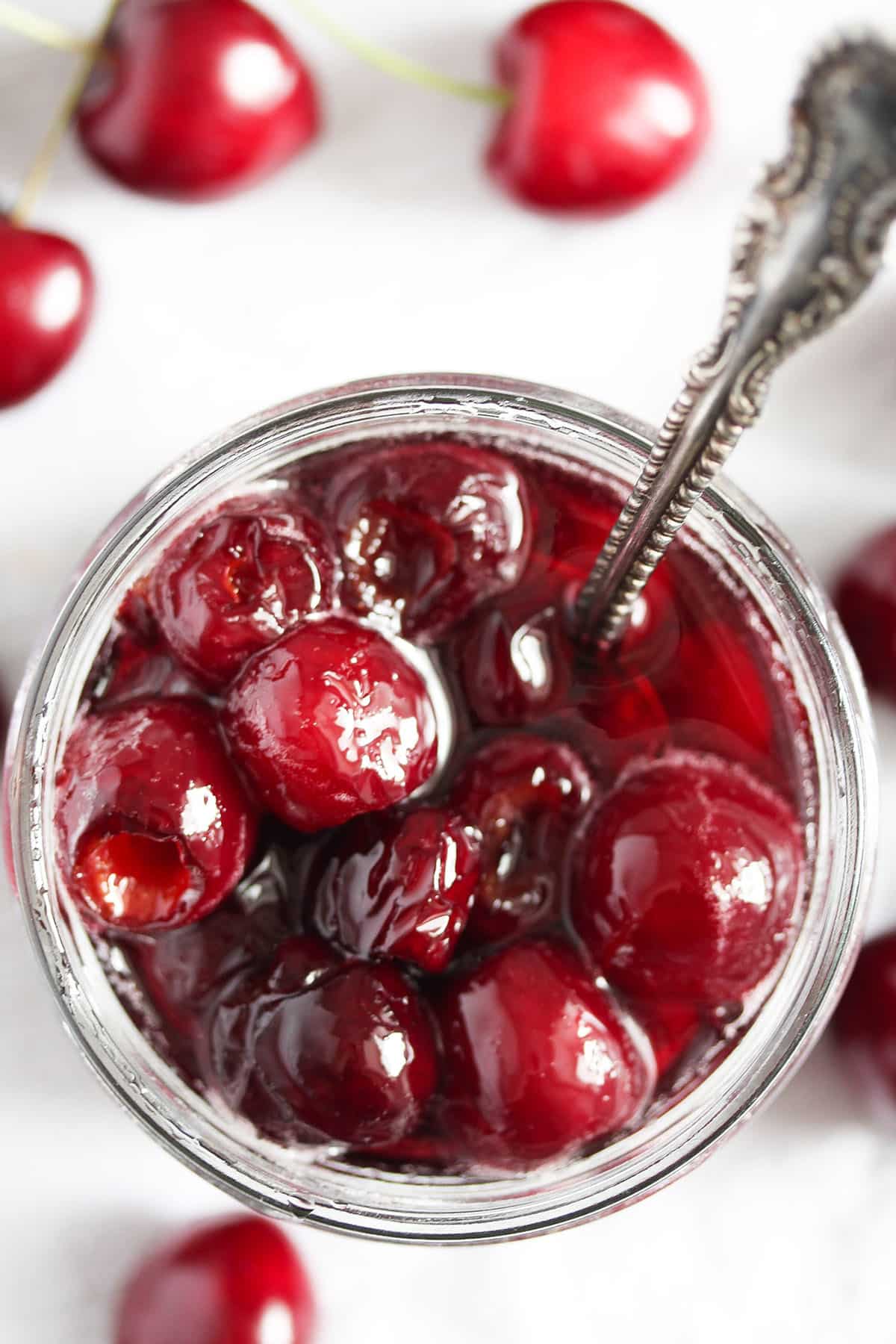 a jar with syrup cherries with a spoon in it and some cherries around it.