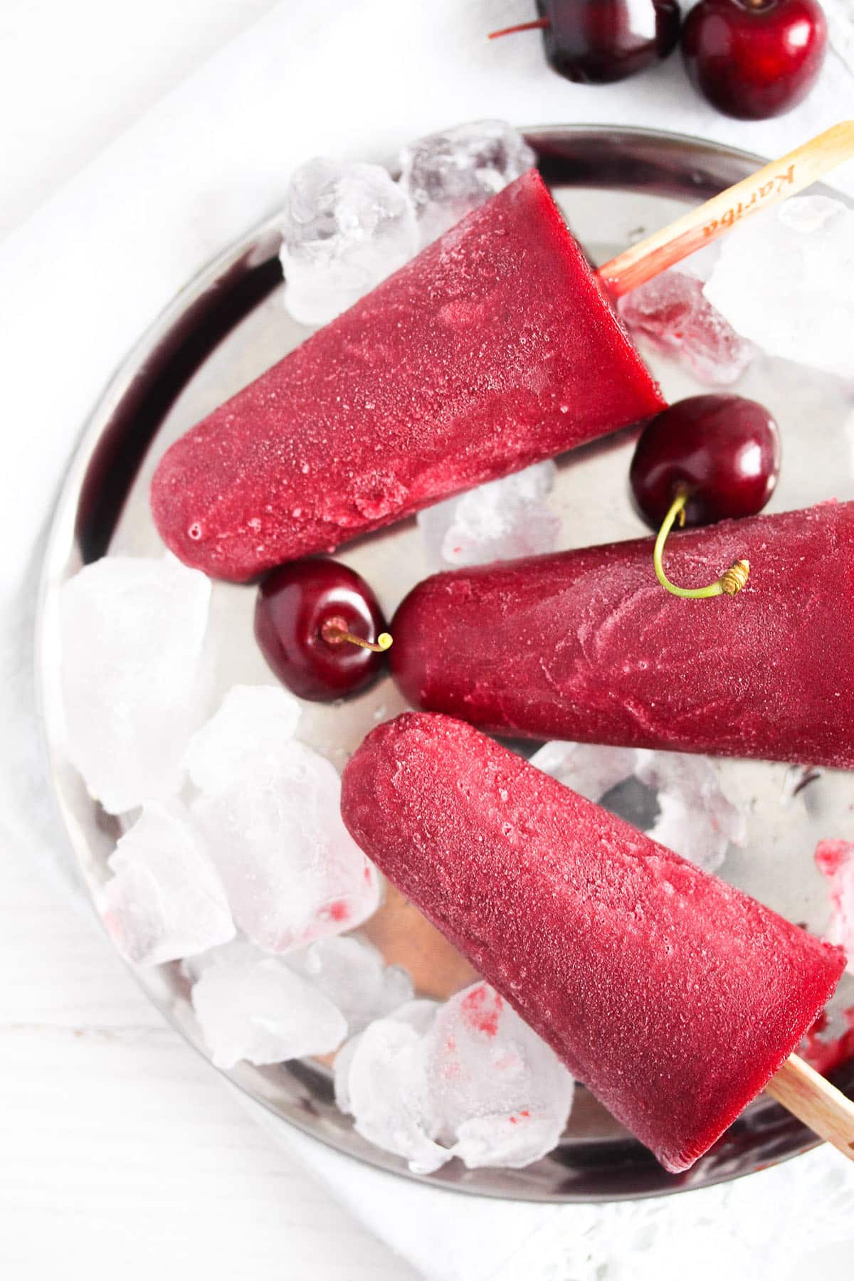 three red cherry popsicles and two cherries on a platter full of ice cubes.