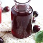 homemade syrup with cherries in a small bottle with cherries behind it.