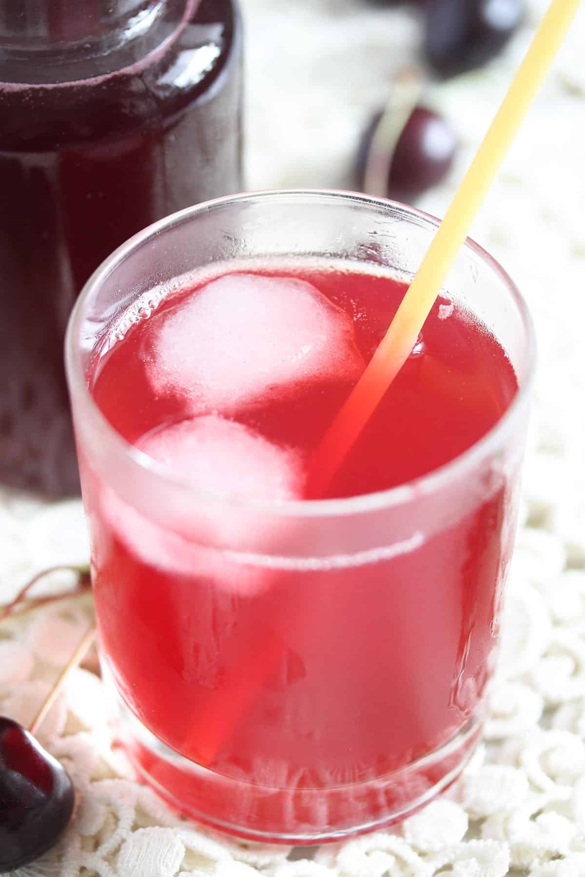 a glass with mixed cherry syrup with sparkling water with two ice cubes and a straw inside.