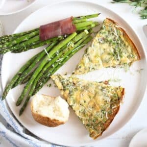 two slices of pea and leek fritatta, half a bun and asparagus with bacon on a white plate.