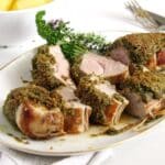 pistachio crusted pork tenderloin sliced on a serving platter with some herbs behind it.