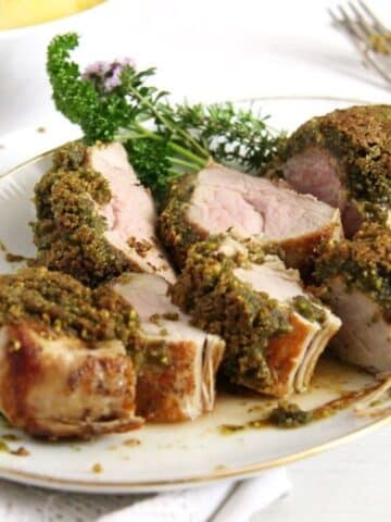 pistachio crusted pork tenderloin sliced on a serving platter with some herbs behind it.