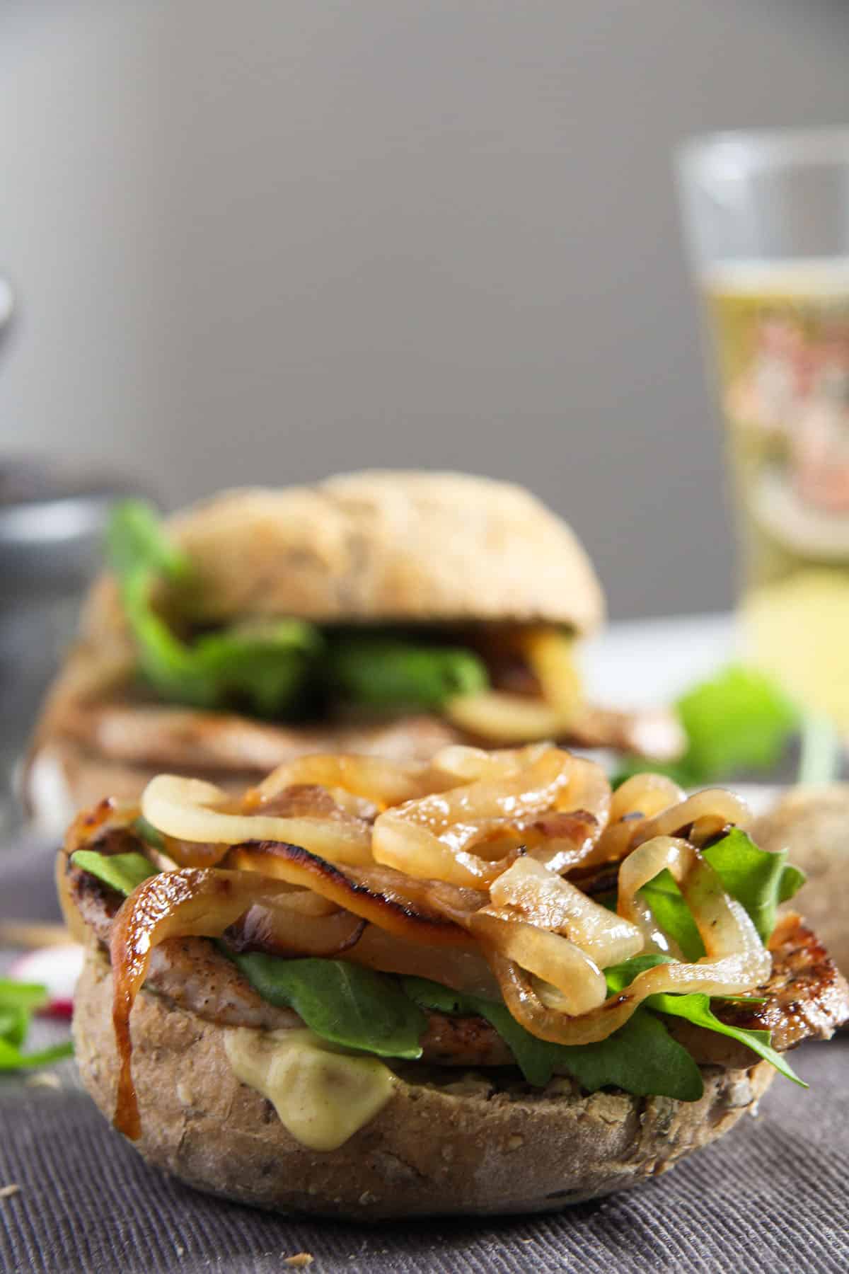 one pork chop burger topped with onionis and arugula, a glass of beer behind it.